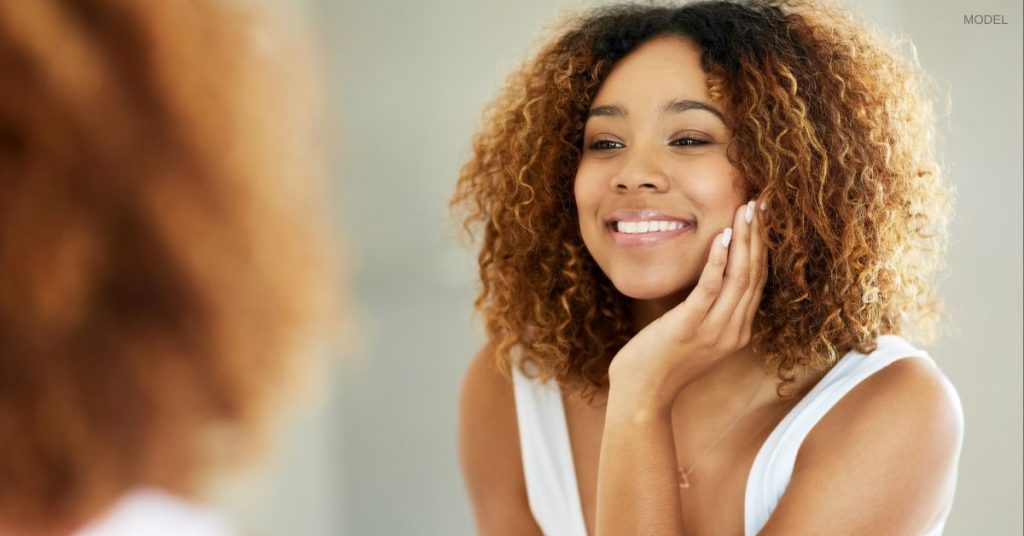 Woman with clear and radiant skin (model) holds a hand up to her face while looking in the mirror.